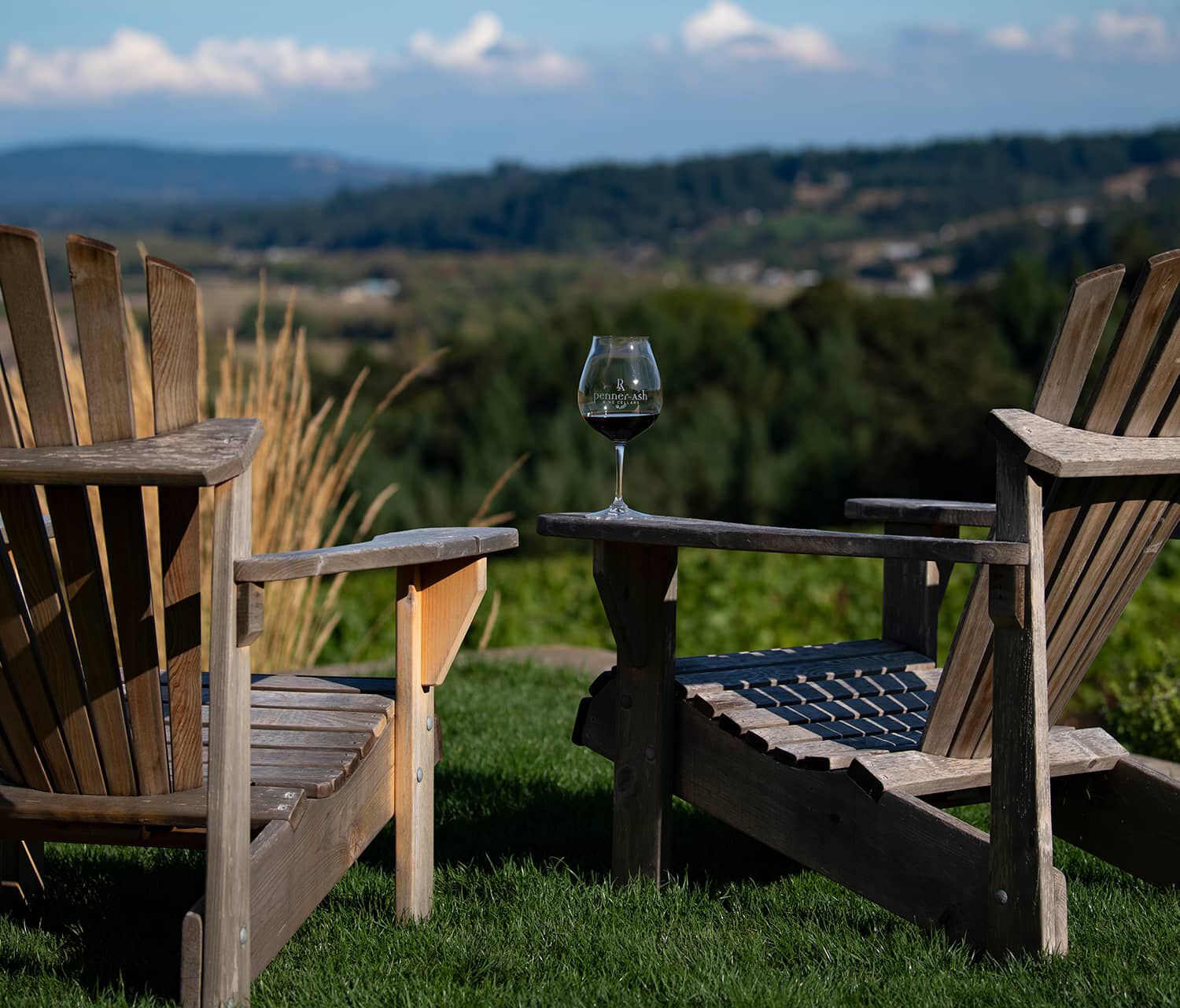 Two Adirondacks with a glass of wine sitting on arm and the Penner-Ash Estate Vineyard in the background.