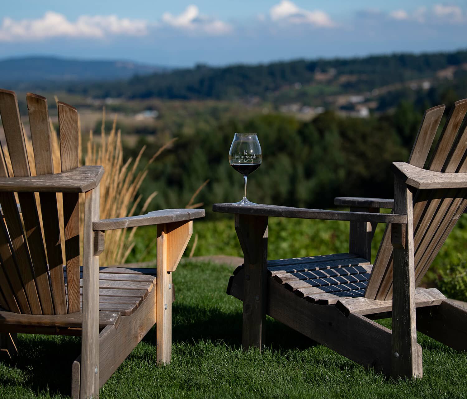 Two Adirondacks with a glass of wine sitting on arm and the Penner-Ash Estate Vineyard in the background. Two Adirondacks with a glass of wine sitting on arm and the Penner-Ash Estate Vineyard in the background.