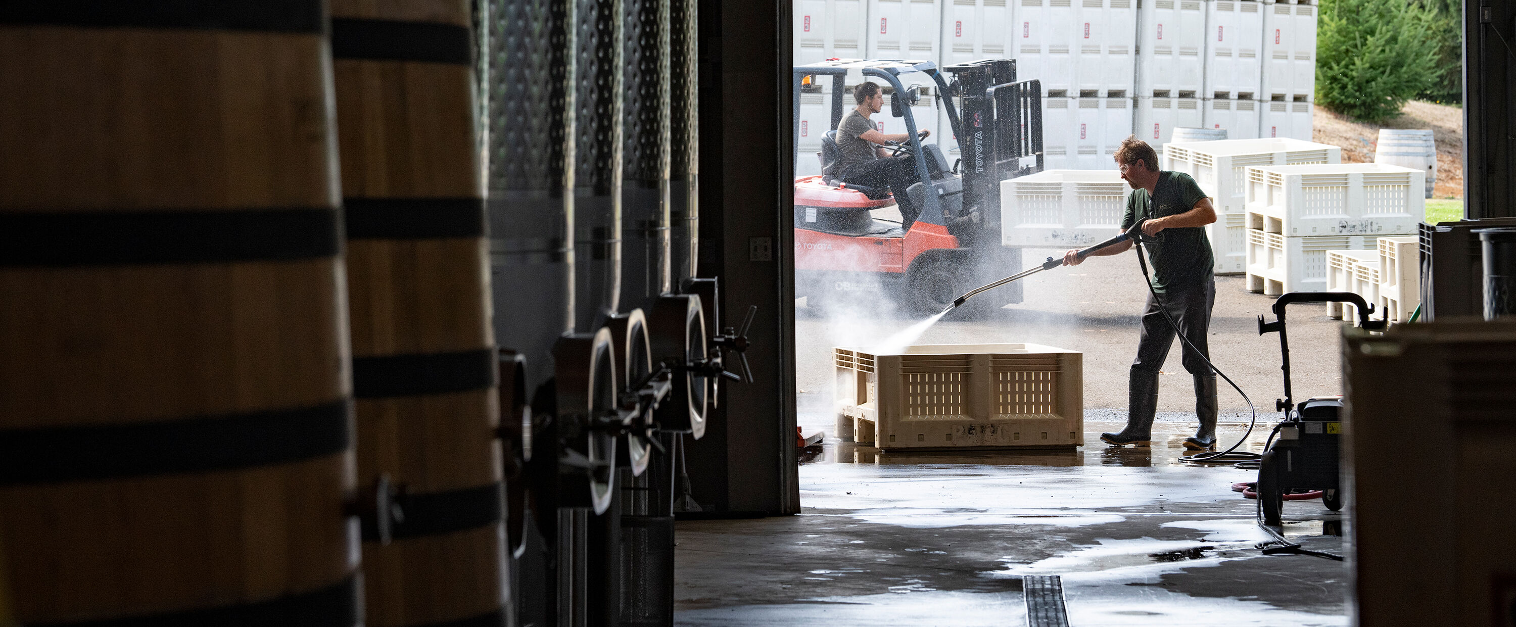 A person cleaning a grape bin with fermentation tanks in the foreground.